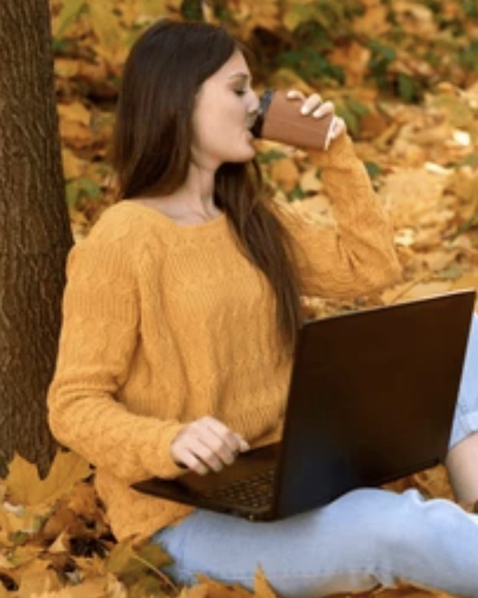 A woman in an amber sweater with a laptop and a coffee, sitting under a tree in autumn leaves